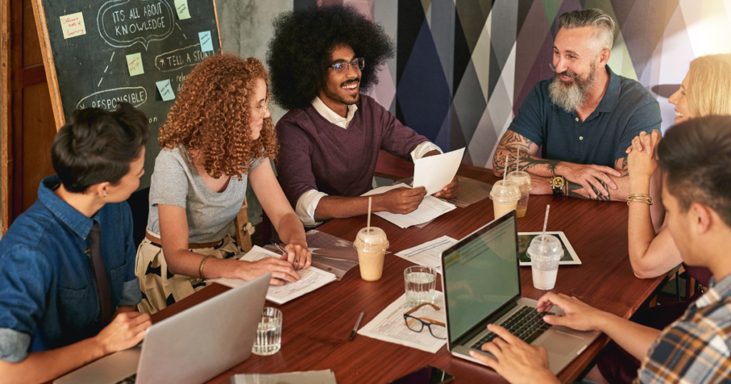 Group of 6 individuals having a meeting with laptops and drinks on the table.